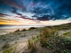 Dramatic landscape view of sand dunes overlooking a beach and ocean under a cloud-filled sky colored in blue and orange.