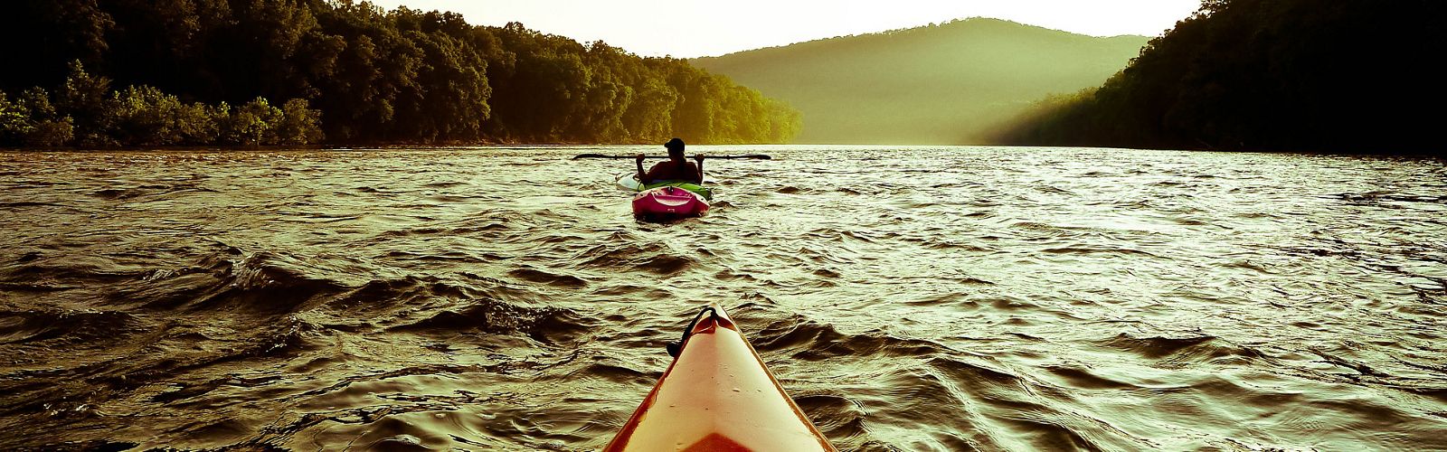 View from a kayak of another kayak on a large body of water with dense forests on all sides.