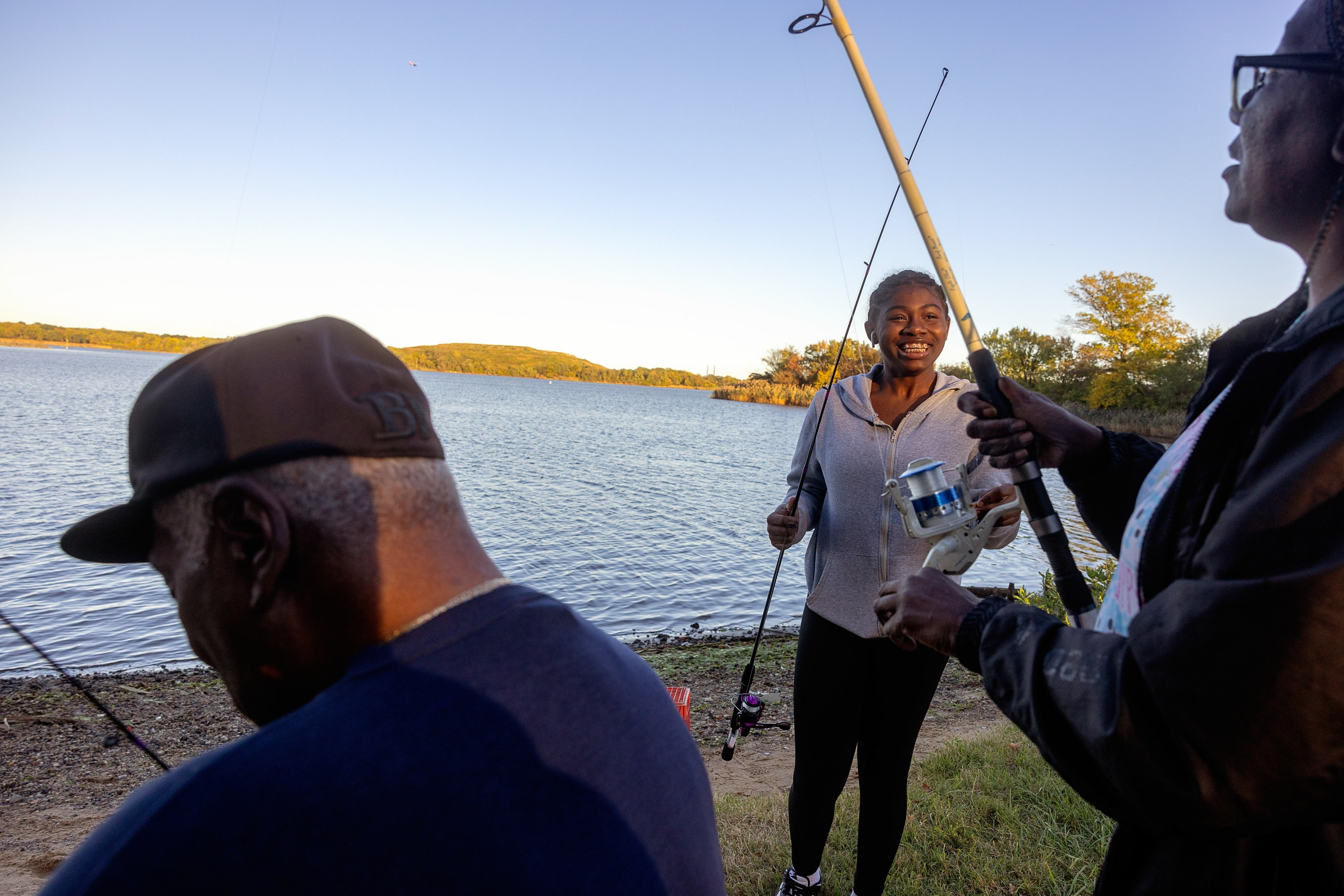 a younger girl holds a fishing pole next to a woman and a man who face the water line.