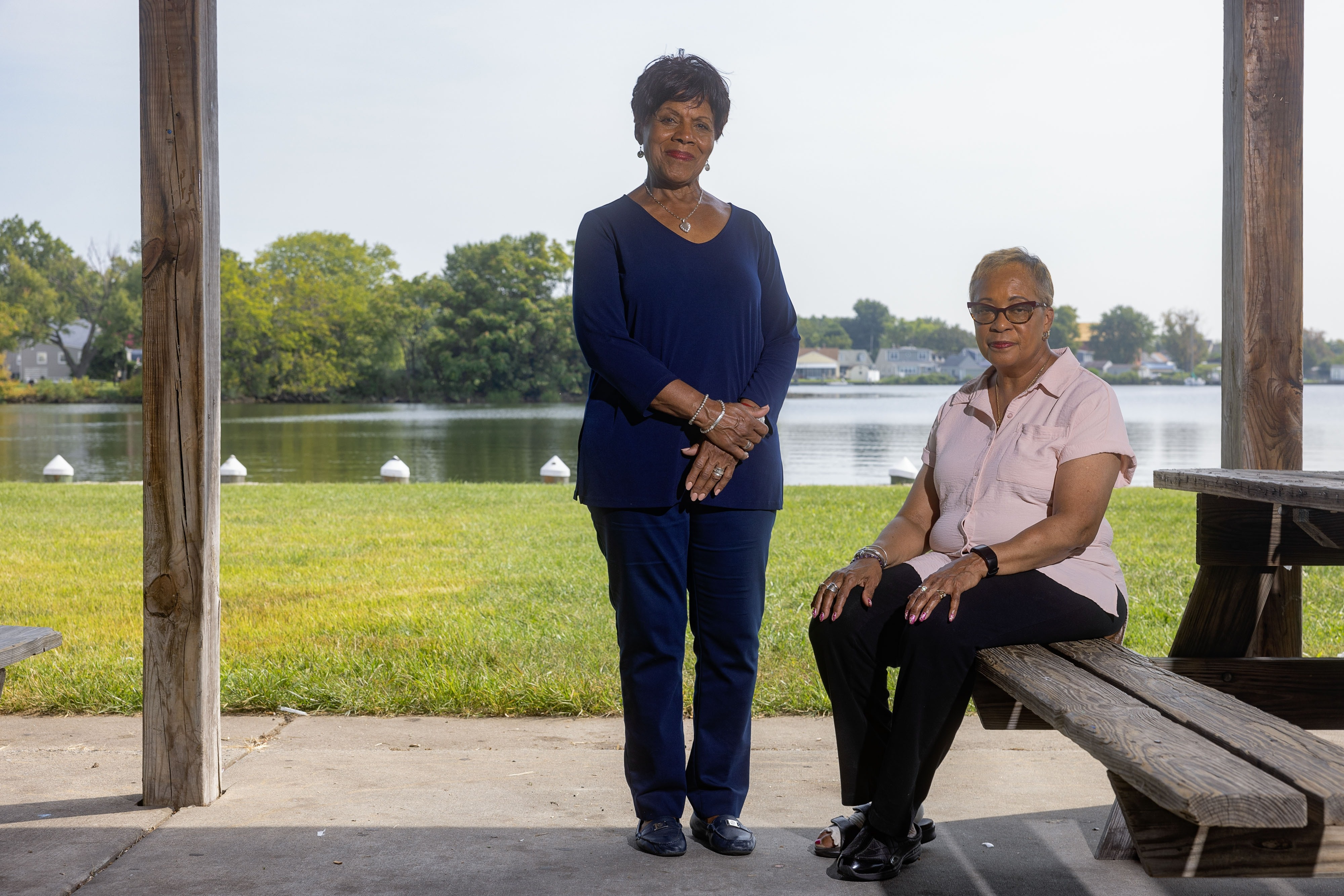 A woman stands beside a woman sitting on a bench in a grassy park along a body of water.