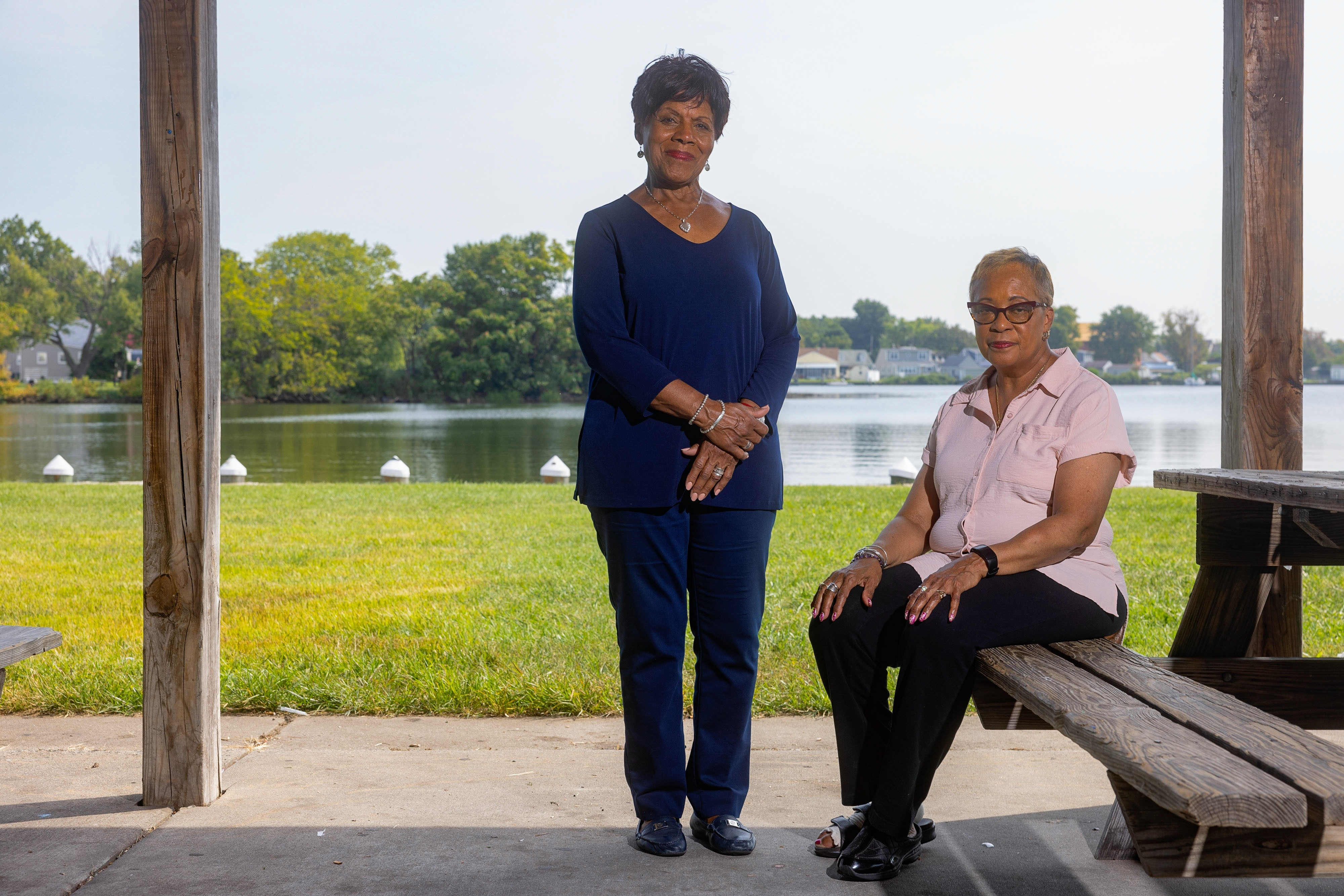 A woman stands beside a woman sitting on a bench in a grassy park along a body of water.