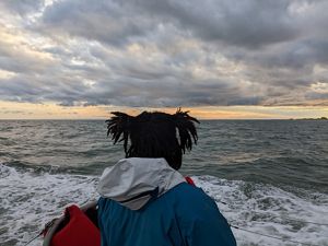 A woman stands on the deck of a boat, facing away from the camera and looking towards the low heavy clouds gathered on the horizon. 